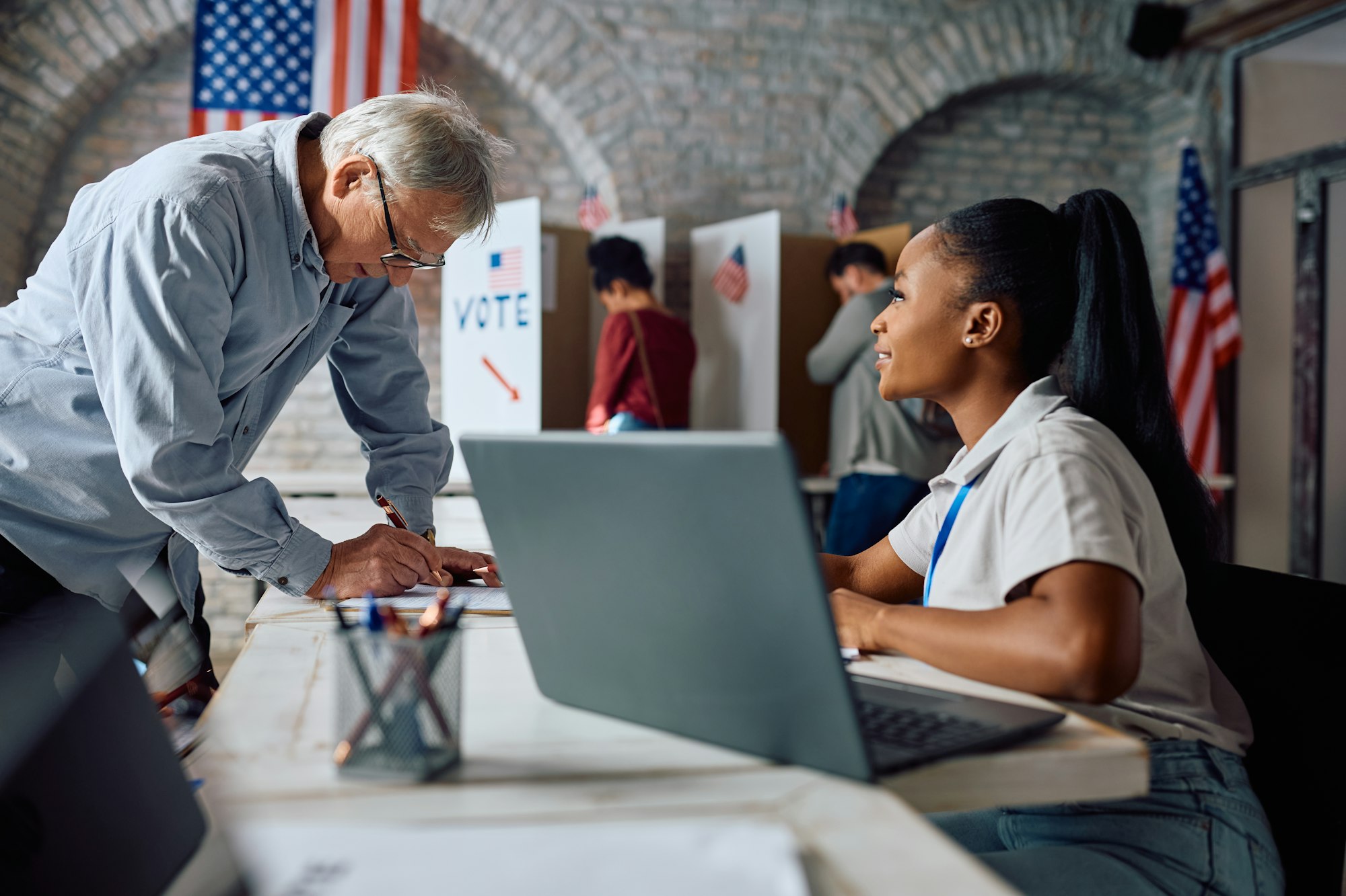 Senior US citizen signing up for voting with help of black female volunteer at polling place.